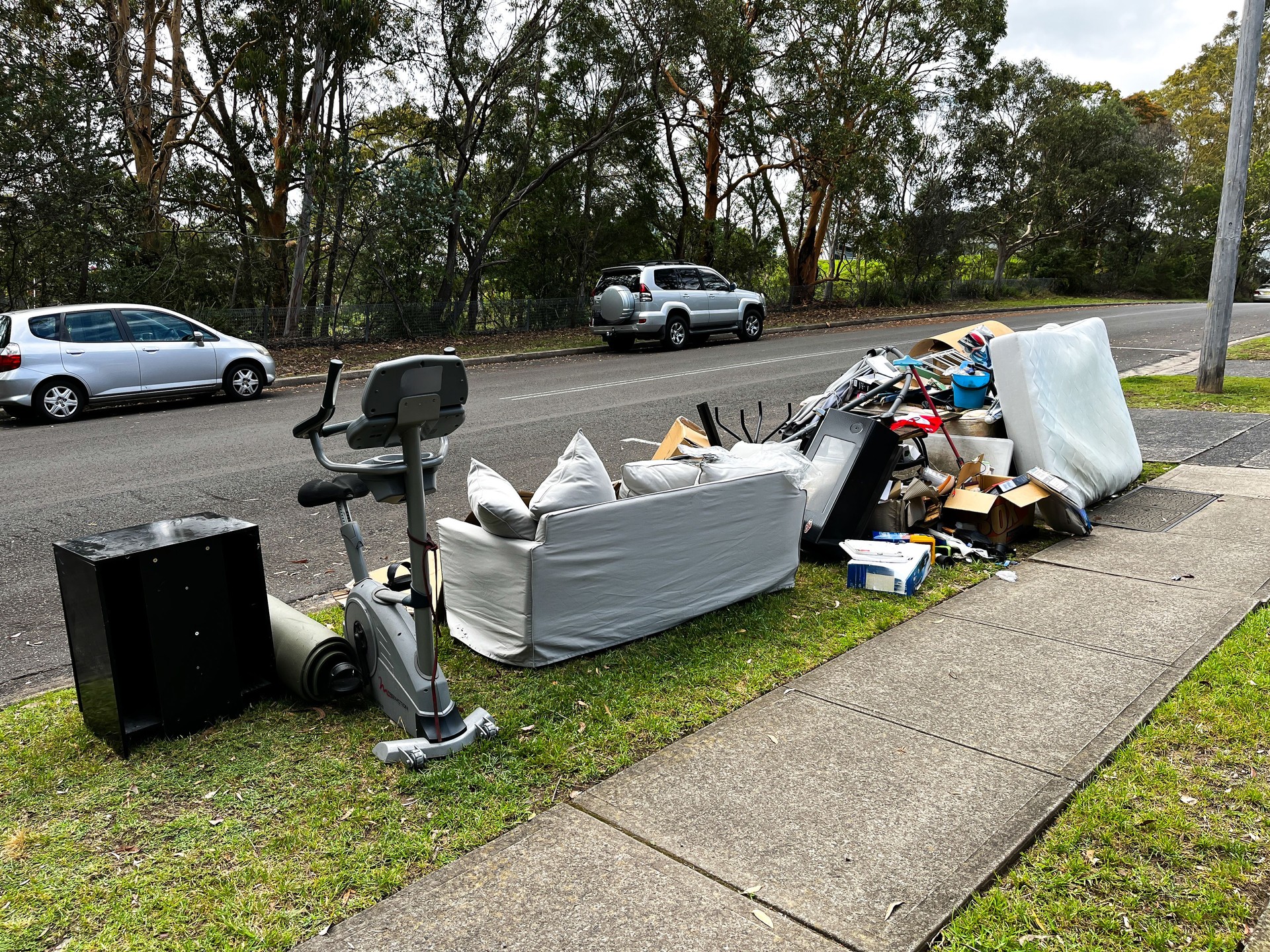 A pile of rubbish on the curb for council collection, including cardboard boxes, a couch with pillows, stacked mattresses, and coat hangers